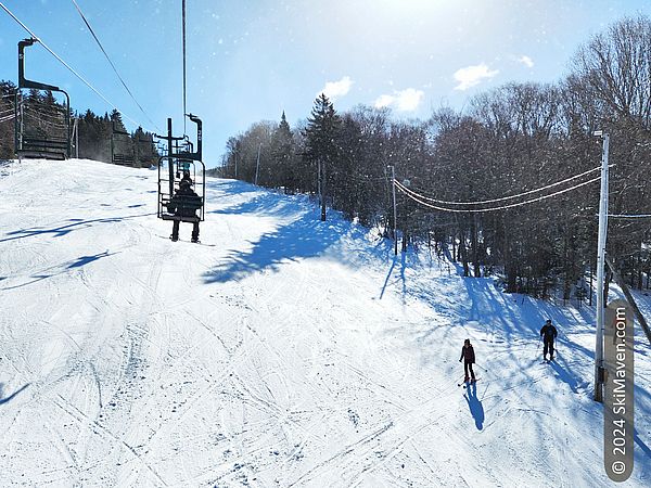 Skiers ride up a double chairlift in sun with skiers below