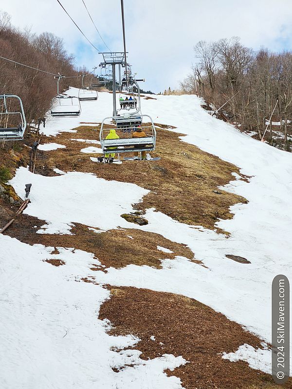 Two skiers ride a quad chairlift over partly grassy ski slope