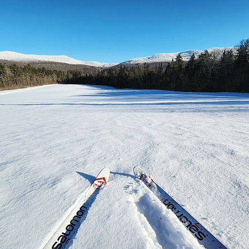 Cross-country skis in foreground at top of snowy hill with snowy mountain range