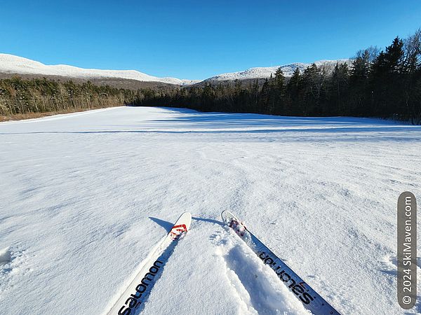 Cross-country skis in foreground at top of snowy hill with snowy mountain range