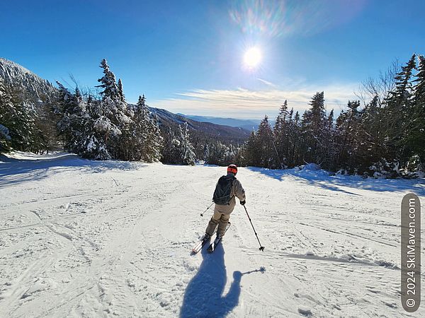 Skier moves downhill at center of ski slope under bright sun