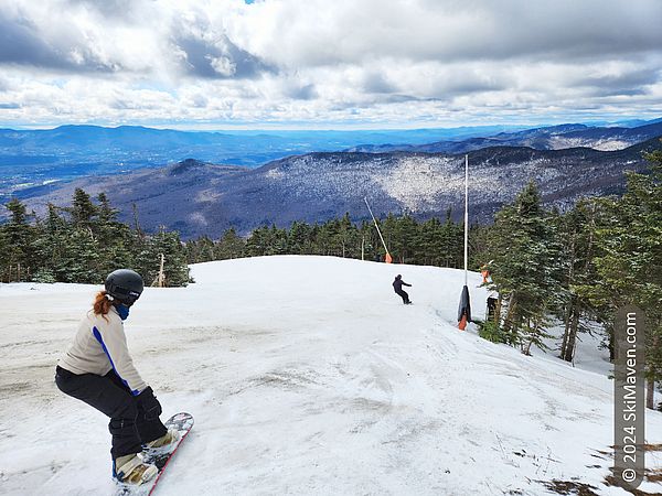 Two snowboarders glide down a slope with mountain views and partly sunny skies