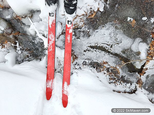 Looking down at a pair of red skis crossing a partially frozen stream