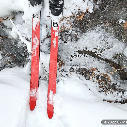 Looking down at a pair of red skis crossing a partially frozen stream