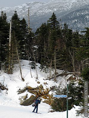 A pastor skis downhill with his guitar.